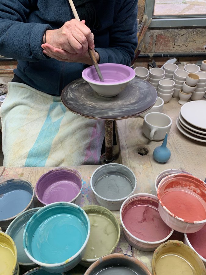 A potter brushing colored glaze onto the interior of a bowl on a banding wheel, surrounded by bowls of different colored glazes