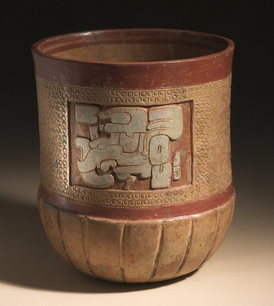 A Maya ceramic vessel with a deep relief-carved panel depicting deity heads, plus carved fluting around its base