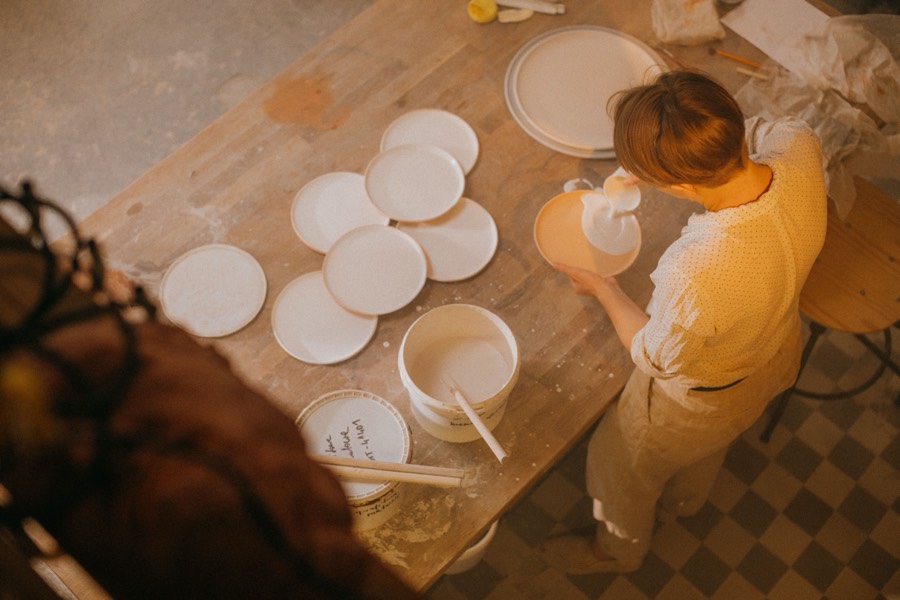 Overhead view of a potter holding a freshly glazed, dripping plate beside a bucket of white glaze and a row of unglazed bisque plates
