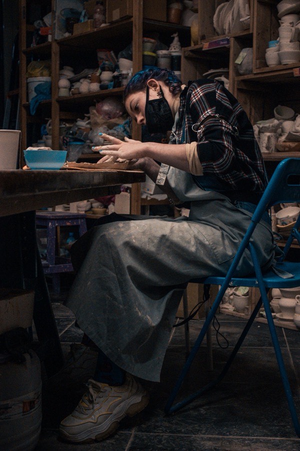 A potter wearing a protective dust mask while working at a table in a ceramics studio, glazed pottery visible on shelves behind them