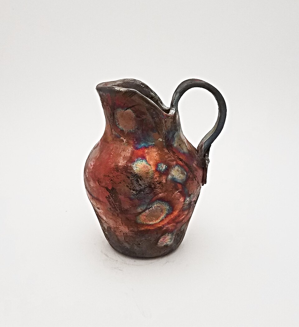 A small ceramic pitcher with an iridescent copper-matte raku glaze in red, blue, and metallic tones, smoke-blackened at the rim and base