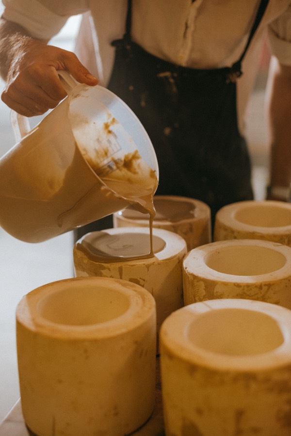 A hand pouring tan liquid slip from a jug into a row of small cylindrical plaster molds
