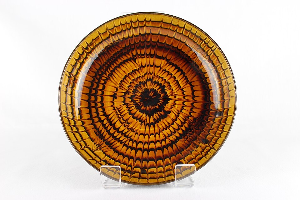A round ceramic dish with a concentric brown and amber feathered slip-trailed pattern radiating from its center