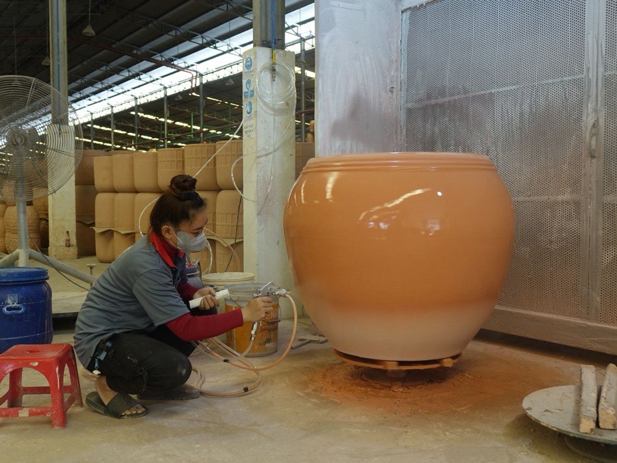 A worker wearing a dust mask kneels beside a large orange-glazed ceramic pot in a workshop, spray-gun in hand, applying glaze to the lower half