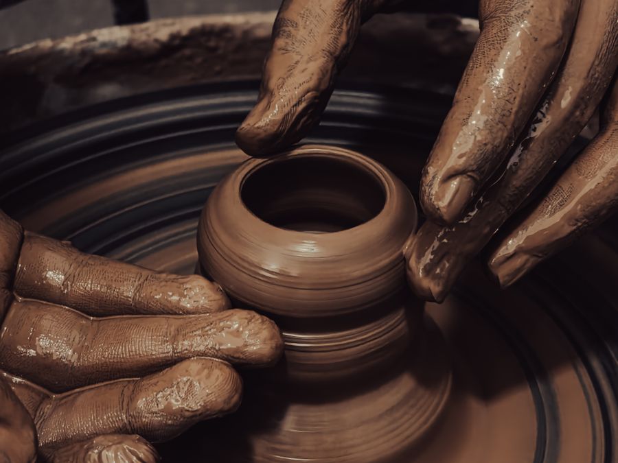 Close-up of clay-covered hands centering and opening a small reddish-brown pot on a spinning potter's wheel